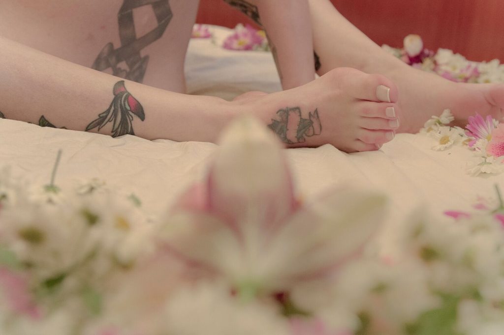 pé tatuado com unhas decoradas estilo "francesinha" e há flores desfocadas no primeiro plano da imagem