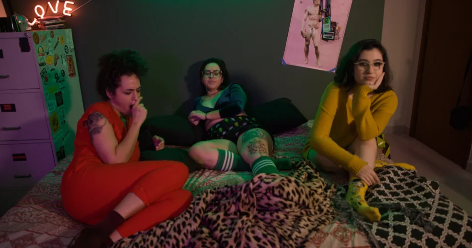 Três amigas sentadas na cama num quarto com neon LOVE