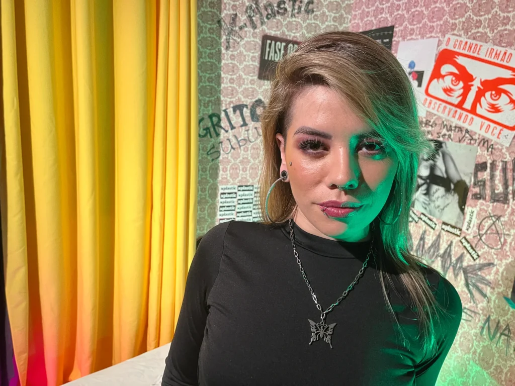 Mulher de blusa preta num quarto com cortina amarela e pôsteres