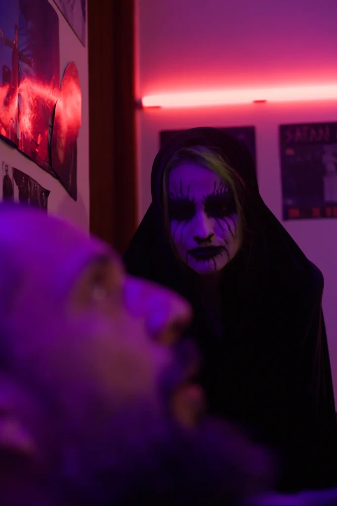 Vlad (Mário) olha assustado para a Entidade (Lady Milf), que surge de capuz preto e corpse paint no quarto com luz neon rosa
