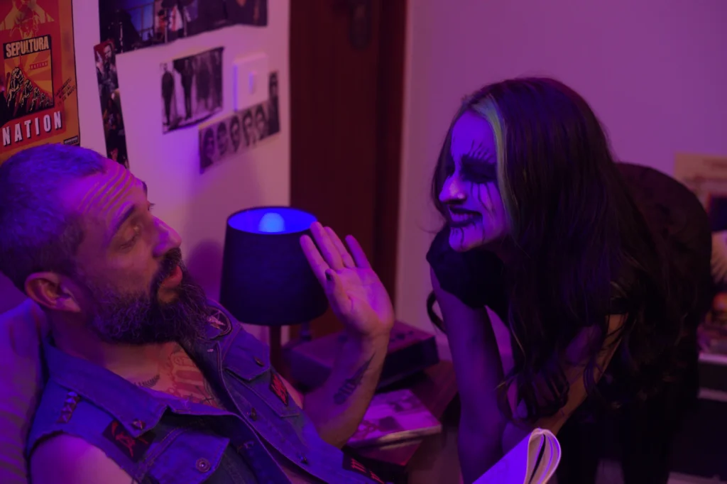 Vlad (Mário) gesticula sentado na cama enquanto a Entidade (Lady Milf) se debruça sorrindo ao lado dele, em cena de Invocação