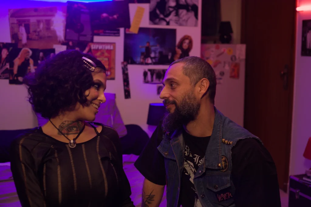 Vlad (Mário), de colete jeans, frente a frente com Maya (Qween) no quarto iluminado de roxo, em cena de Invocação