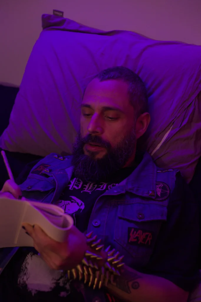 Vlad (Mário) deitado na cama com colete jeans cheio de patches de bandas de metal, escrevendo num caderno, em cena de Invocação