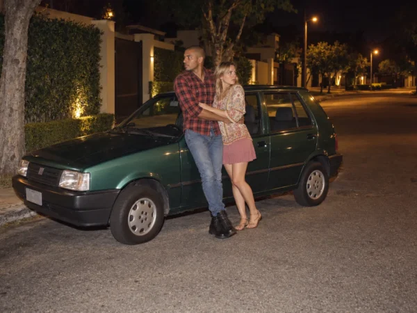 Casal abraçado encostado num carro numa rua residencial à noite, olhando em volta